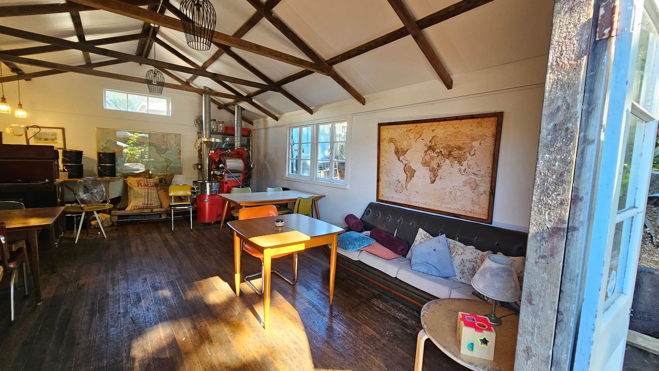 A rustic interior with wooden beams on the ceiling and a wooden floor. On the right, a sofa with assorted cushions is placed against the wall, above which hangs a large world map. A small table with a lamp and a toy is nearby. In the centre, a wooden table with chairs is illuminated by sunlight. On the left, there are more tables, chairs, and a coffee roaster. The walls have windows and framed pictures, and the space is lit by hanging lights.