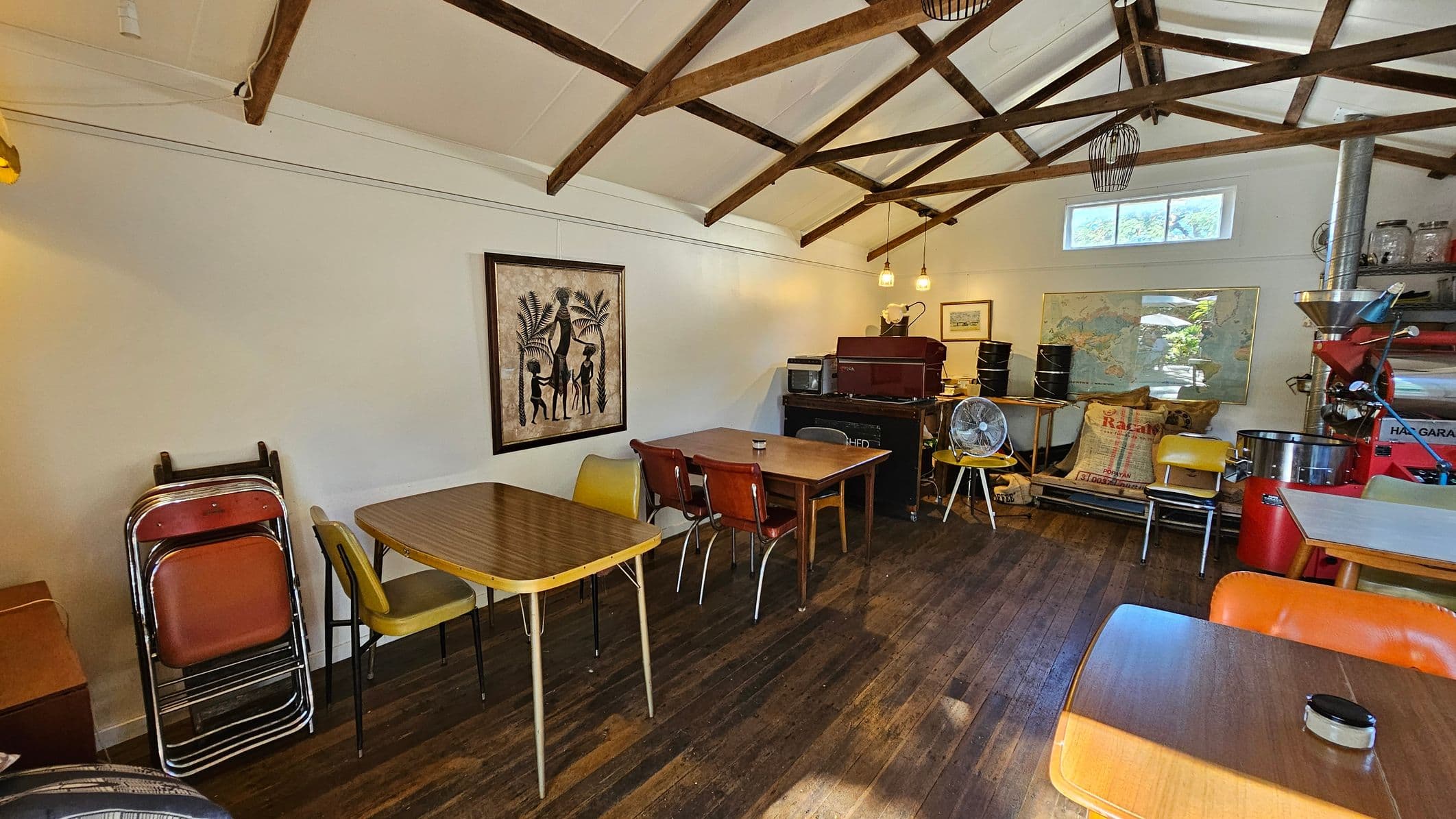A cosy room with a wooden floor and exposed wooden beam ceiling. On the left, there are stacked chairs and a table with mismatched chairs. A framed artwork depicting figures and palm trees hangs on the wall. In the centre, more tables and chairs are arranged, with a small fan on one table. The right side features a coffee roaster, bags of coffee beans, and a map on the wall. Pendant lights hang from the ceiling, and a small window lets in natural light.