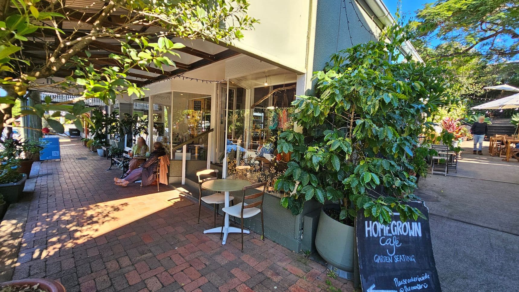 A café with outdoor seating is on the right, featuring a chalkboard sign that reads "Homegrown Café Garden Seating." Potted plants and a small table with chairs are nearby. On the left, a brick pathway is lined with more plants and a few people sitting on benches. The café has large windows, and the area is shaded by a wooden pergola. In the background, there are more tables with umbrellas and a person walking away.