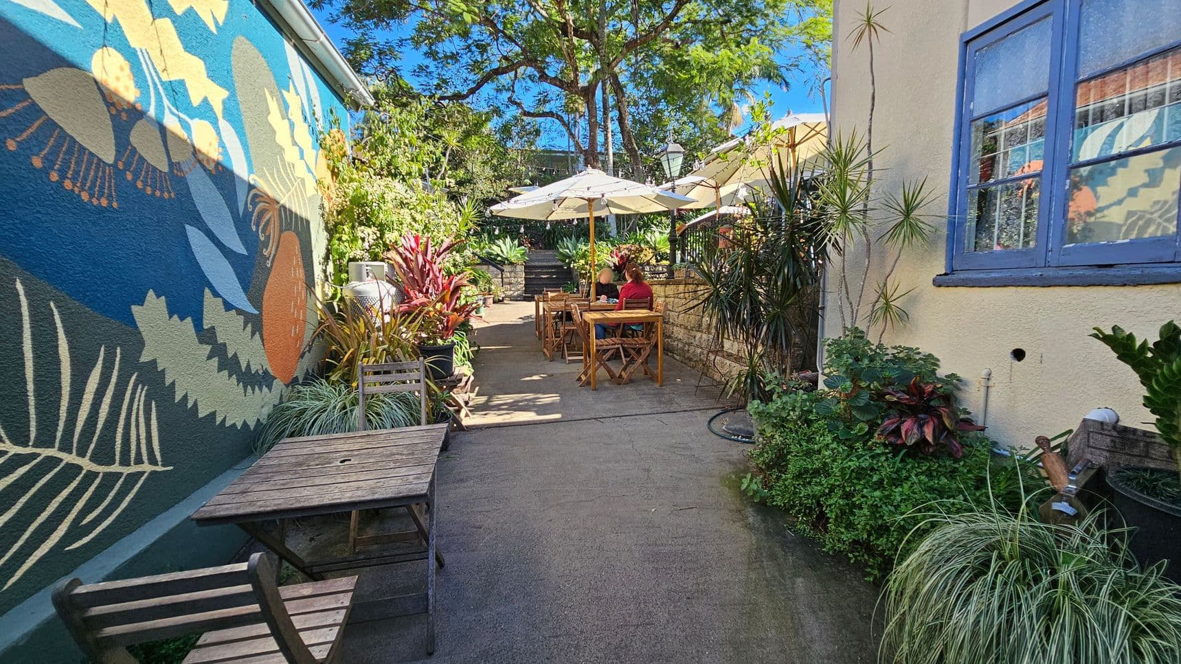 A narrow outdoor café area with a colourful mural on the left wall featuring abstract plant designs. Wooden tables and chairs are placed along the path, with a couple seated at one table under large white umbrellas. Lush greenery and potted plants line both sides, creating a vibrant atmosphere. The right side has a light-coloured wall with a blue-framed window. In the background, a tree provides shade, and steps lead to another area. The ground is paved with concrete.