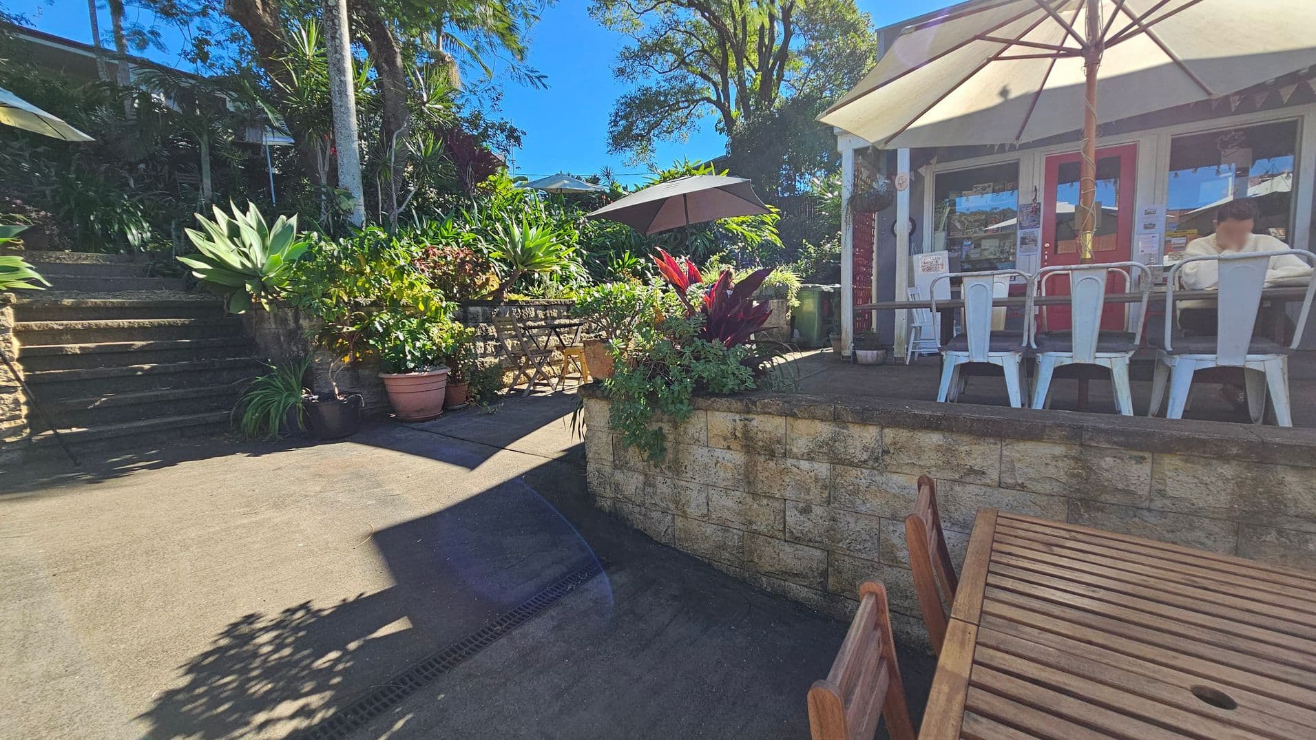 Outdoor café setting with a wooden table and chairs in the foreground. On the right, a café with a red door and white-framed windows, featuring metal chairs and a large umbrella. A person is seated at a table. In the middle, a stone wall with lush green plants and red foliage. On the left, stone steps lead up to more greenery and a shaded area with additional tables and chairs. Tall trees and a clear blue sky are visible in the background.