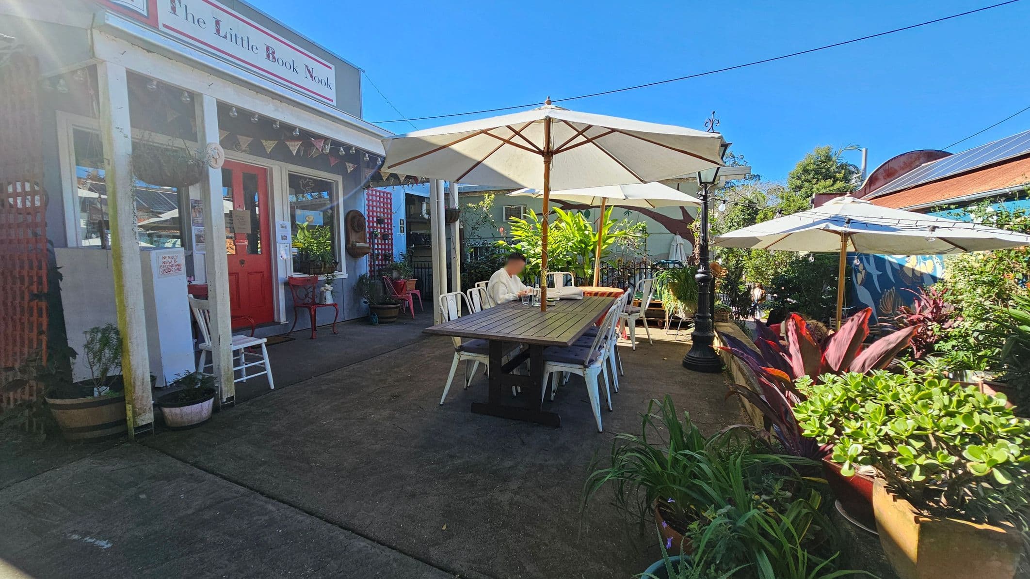 A quaint outdoor seating area with a long wooden table and white chairs under large white umbrellas. On the left, a building with a red door and a sign reading "The Little Book Nook" is adorned with bunting. Potted plants and greenery surround the area, adding a lush feel. A person is seated at the table, and a lamppost stands nearby. The ground is paved, and the background features a mural and more plants.