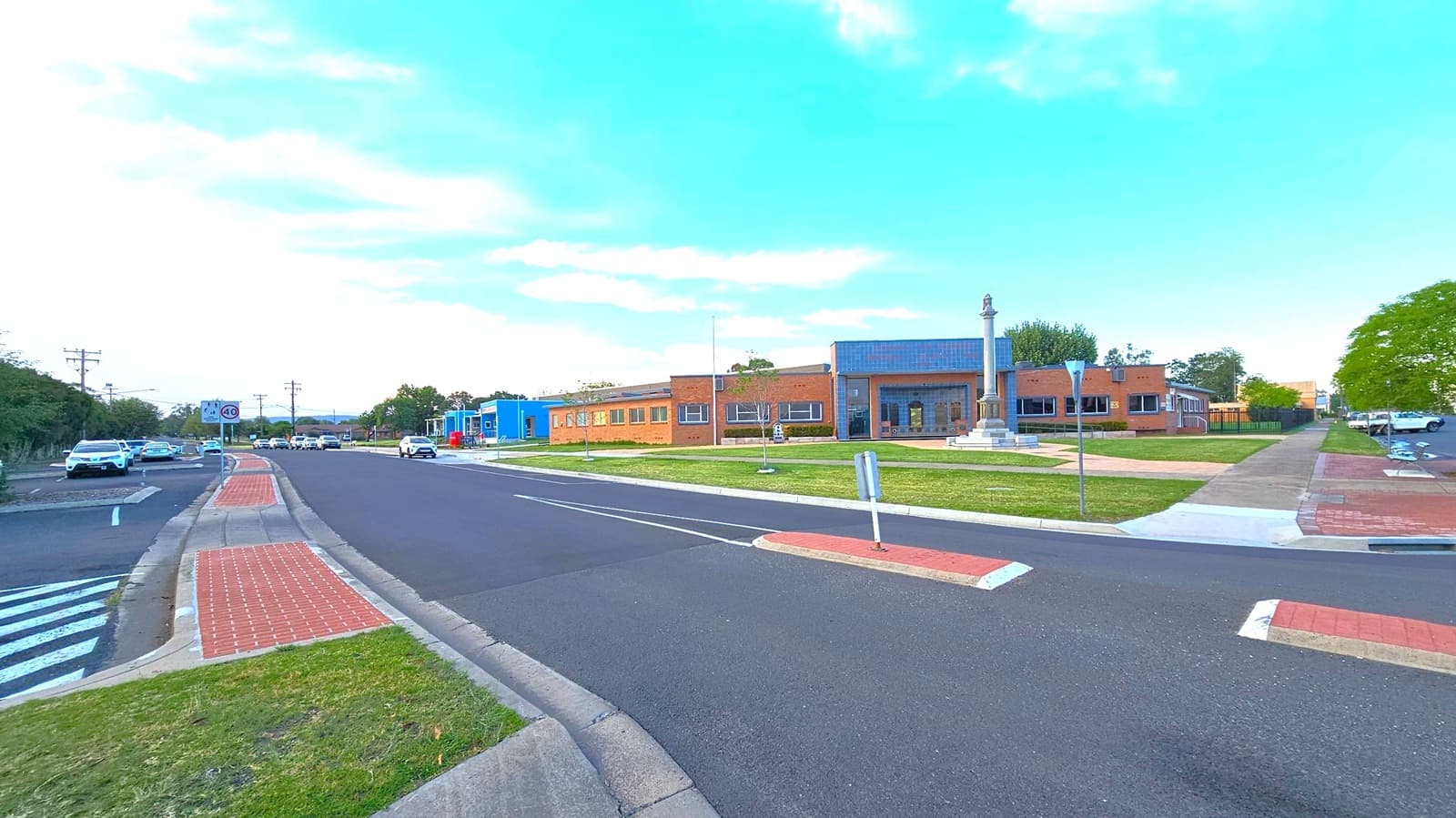 A wide street with a central median and a pedestrian crossing. On the left, several cars are parked along the road, with a speed limit sign indicating 40. On the right, a grassy area with a monument featuring a tall column. Behind it, a series of brick buildings with large windows. The pavement is a mix of red bricks and concrete, and there are trees in the background. The sky is clear with some clouds.