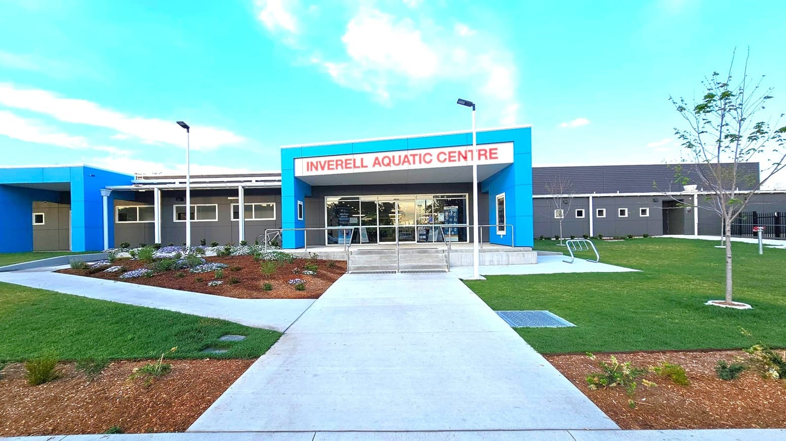 A modern building with a blue and grey facade, featuring a sign that reads "Inverell Aquatic Centre" in red letters above the entrance. The entrance has glass doors and is flanked by two white lamp posts. In the foreground, a concrete path leads to the entrance, bordered by landscaped gardens with small plants and mulch. On the right, there is a grassy area with young trees. The building has a flat roof and several small windows.