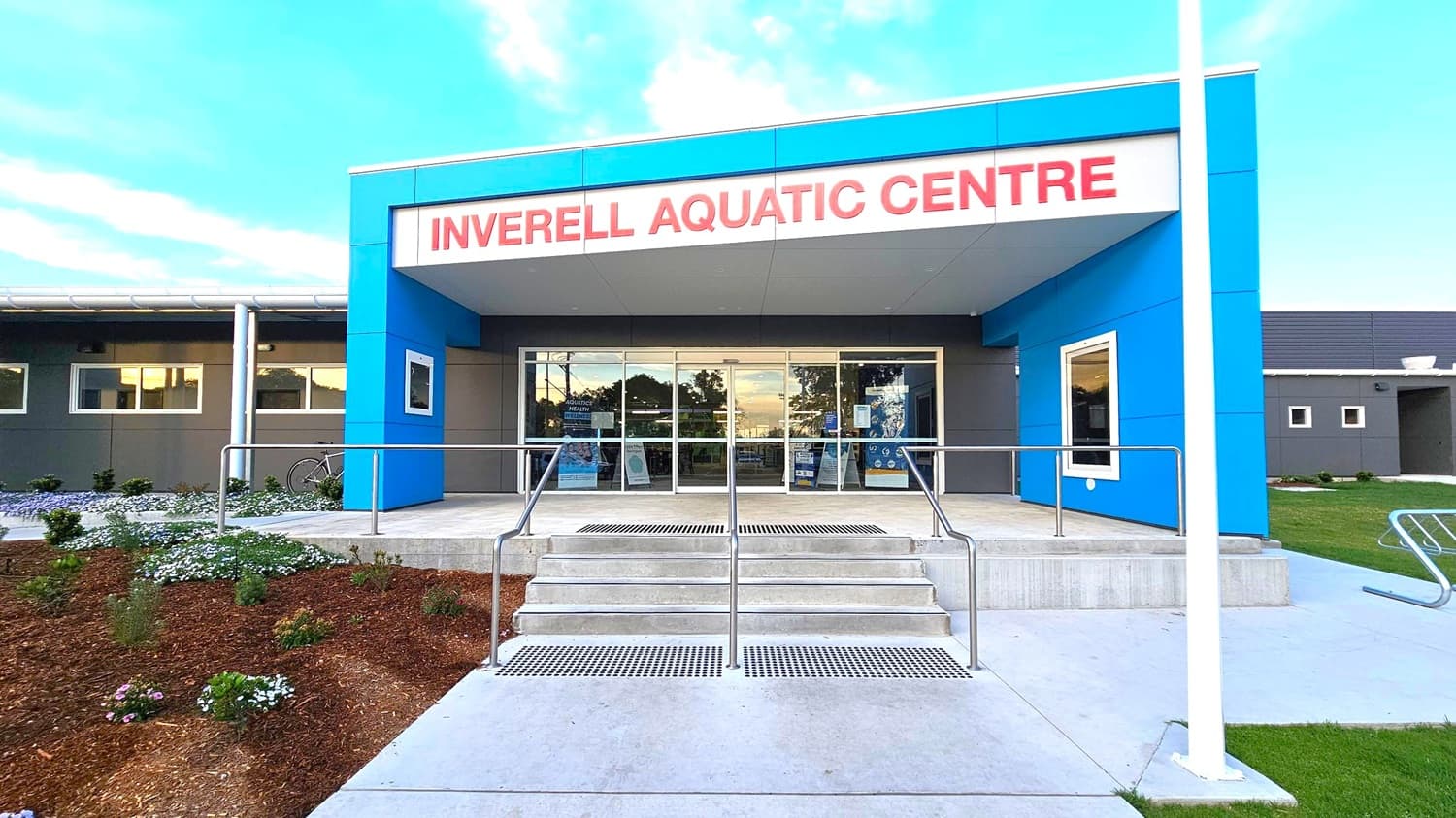 The entrance of the Inverell Aquatic Centre features a modern design with a blue and grey facade. Large glass doors are centrally located, flanked by windows. A set of concrete steps with metal railings leads up to the entrance. On the left, there is a landscaped area with small plants and flowers, and a bicycle is parked nearby. On the right, a bike rack is visible on the concrete path. The building has a flat roof, and the area is well-maintained.
