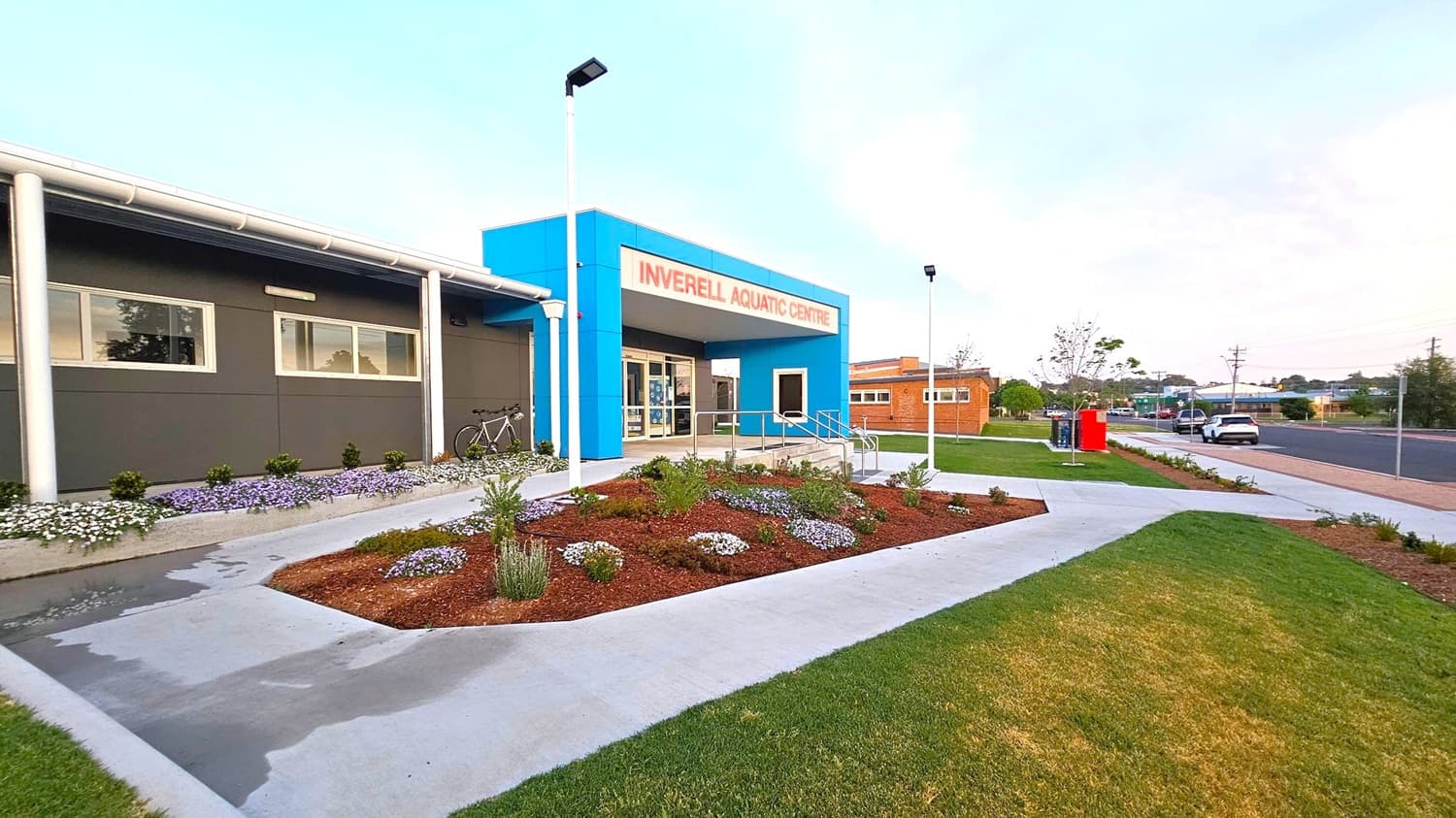 A modern building with a blue entrance labelled "Inverell Aquatic Centre" in red. The left side features a grey wall with windows and a bicycle parked nearby. In front, there is a landscaped garden with various plants and flowers. The right side shows a pathway leading to a street with a parked car and a red utility box. The area is well-lit with street lamps, and the ground is a mix of concrete paths and grassy patches.