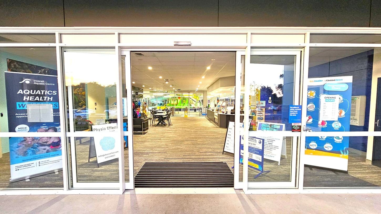 Glass entrance doors to a facility with signs on both sides. On the left, a banner for an aquatic centre and a sign for "Physio Effect." On the right, a poster with opening hours and a progression chart. Inside, a carpeted floor leads to tables and chairs, with a green slide visible in the background. The ceiling is fitted with recessed lighting.