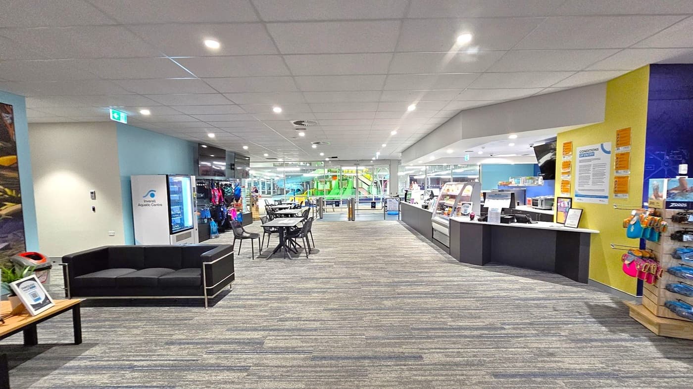 A spacious indoor area with a grey and green carpeted floor and a white panelled ceiling with recessed lighting. On the left, there's a black sofa and a vending machine labelled "Inverell Aquatic Centre." In the centre, tables and chairs are arranged, leading to glass doors revealing a green and blue water slide. On the right, a counter with a display case and a wall with notices and merchandise, including swim gear, is visible.