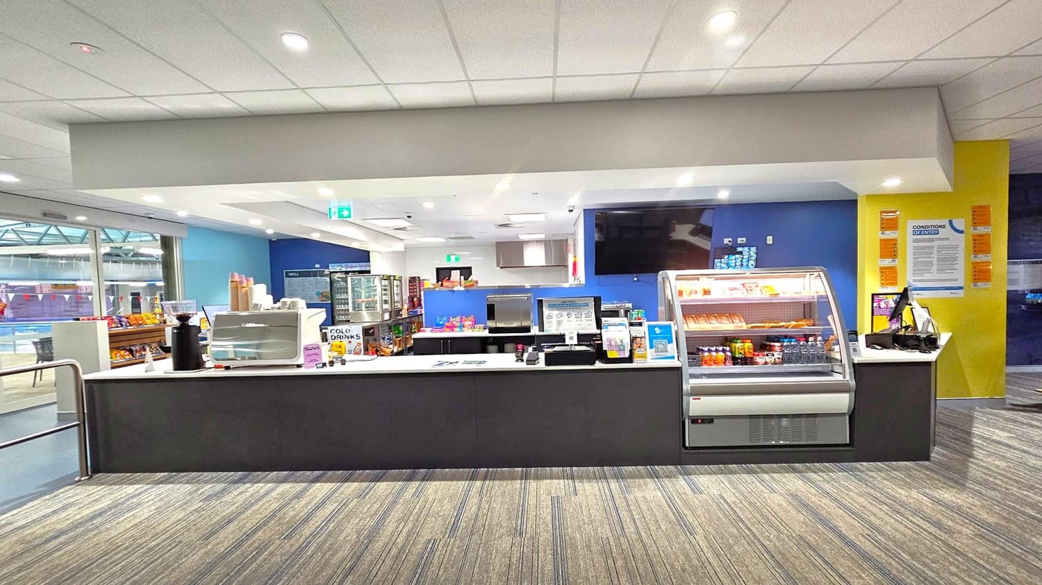 A snack bar with a counter displaying various items. On the left, there are shelves with snacks and a coffee machine. In the centre, a refrigerated display case contains drinks and snacks. Behind the counter, a menu board and a refrigerator with more items are visible. The right side has a yellow wall with informational posters. The floor is carpeted with a striped pattern, and the ceiling is white with recessed lighting.