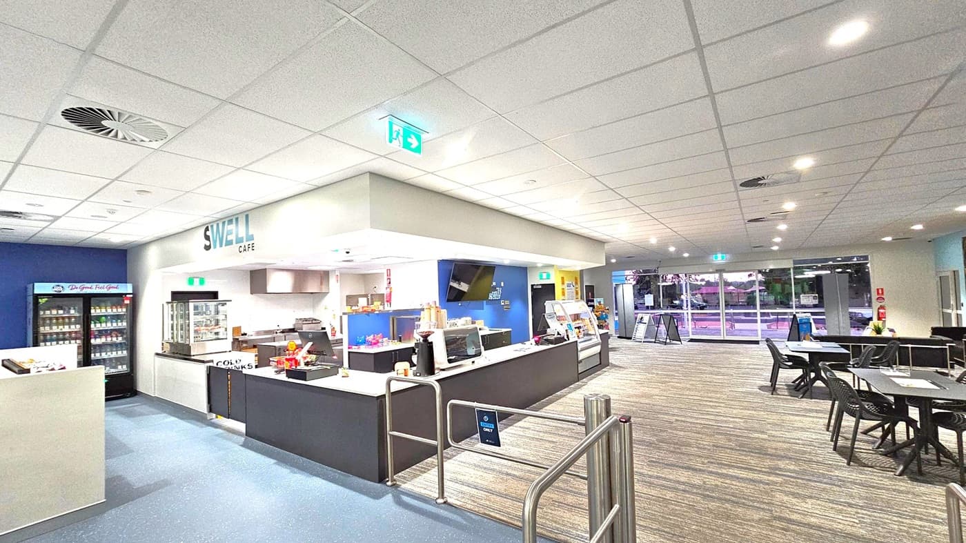 A café interior with a counter in the centre displaying snacks and a coffee machine. The sign above reads "Swell Café". To the left, a refrigerated display holds drinks. On the right, there are tables and chairs on a carpeted floor. The ceiling is fitted with recessed lighting and an exit sign. Large windows in the background show an outdoor view.