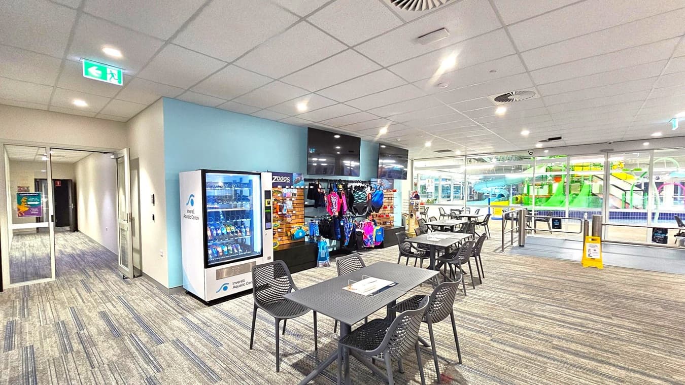 A room with a vending machine on the left, next to a display of swimwear and accessories. In the centre, there are tables and chairs with a magazine on one table. The floor is carpeted with a striped pattern. On the right, large glass windows reveal a view of an indoor pool area with colourful slides. The ceiling is fitted with recessed lighting and air vents. An exit sign is visible above a door on the left.
