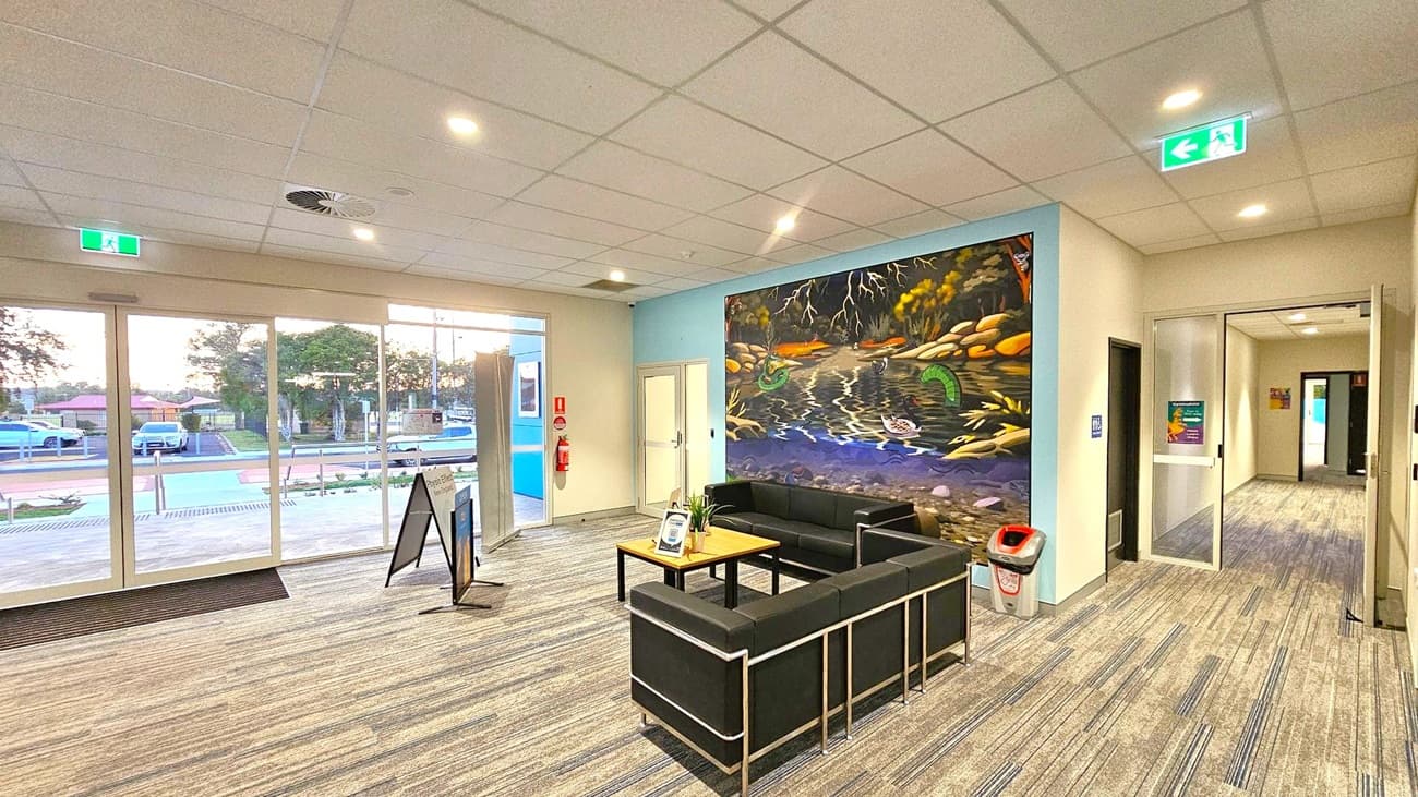 A spacious indoor area with a patterned carpet floor and a grid-patterned ceiling with recessed lights. On the left, large glass doors and windows reveal a view of parked cars and trees outside. In the centre, a seating area with black sofas and a wooden table. A vibrant mural of a river scene with rocks and foliage adorns the wall behind the sofas. On the right, a hallway leads further into the building, and a green exit sign is visible above. A fire extinguisher and a bin are near the mural.