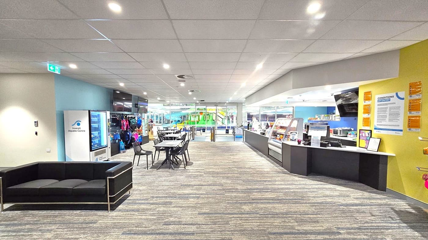 A spacious indoor area with a patterned carpet floor and a tiled ceiling with recessed lighting. On the left, there's a black sofa and a vending machine labelled "Inverell Aquatic Centre." In the centre, several tables and chairs are arranged. On the right, a counter with a display case and various notices on a yellow wall. In the background, large glass windows reveal a view of a pool area with green water slides.