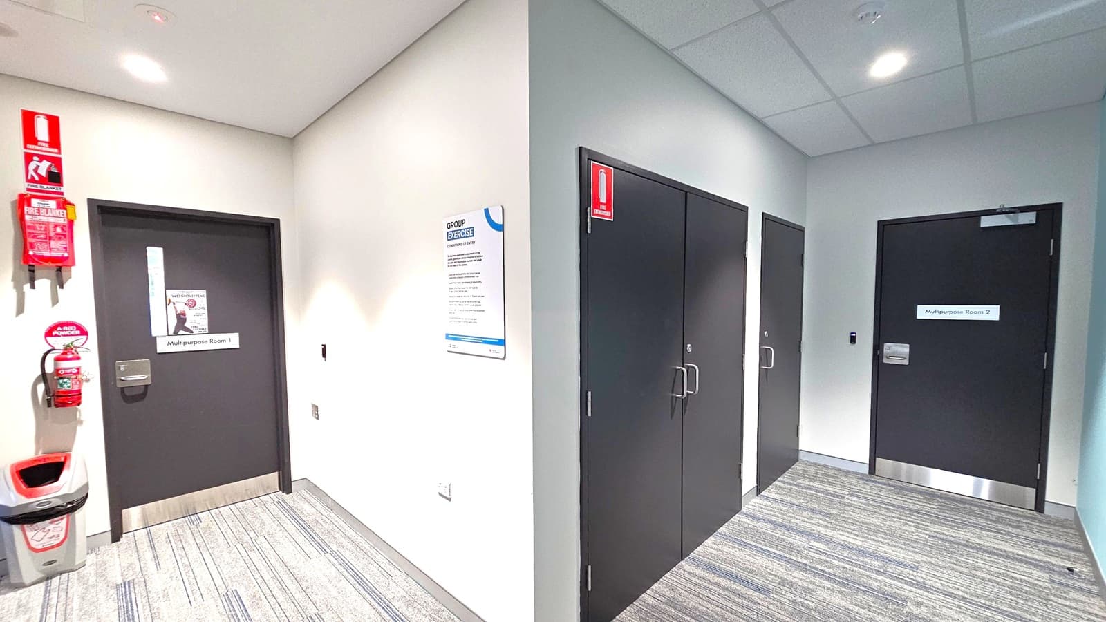 A hallway with grey carpet and a white ceiling featuring recessed lights. On the left, a black door labelled "Multipurpose Room 1" is accompanied by a fire extinguisher, fire blanket, and a bin. A sign about group exercise is mounted on the wall. On the right, two black doors are visible, one labelled "Multipurpose Room 2" and another unlabelled. Both sides have emergency exit signs above the doors.