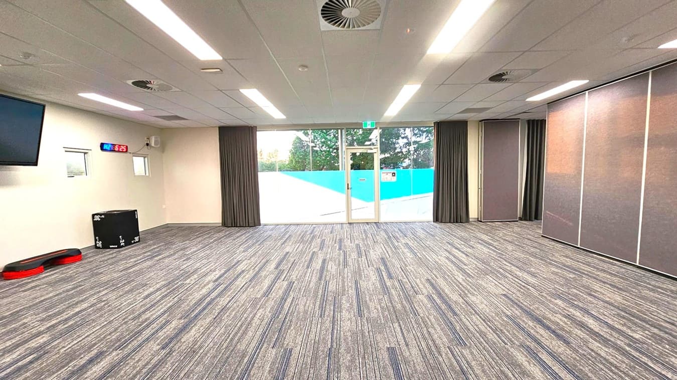 A spacious room with a grey carpet featuring subtle coloured stripes. The ceiling has recessed lighting and air vents. On the left, a wall-mounted digital clock displays "6:25" in red and blue. Below it, a black exercise box and red step platforms are visible. The back wall has large glass doors with a blue and white design, flanked by dark curtains. On the right, there are large, foldable partition panels.