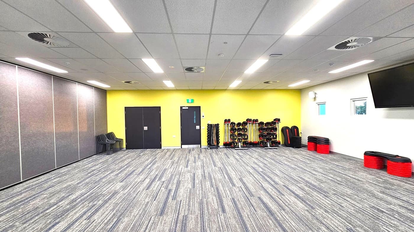 A spacious room with a grey carpeted floor and a ceiling with fluorescent lights and air vents. On the left, there are grey partition walls and a stack of black chairs. The back wall is yellow with two black doors. In the centre, there are racks of weights and exercise equipment. On the right, a white wall features two small windows and a large TV screen. Red and black exercise steps are stacked against the wall.