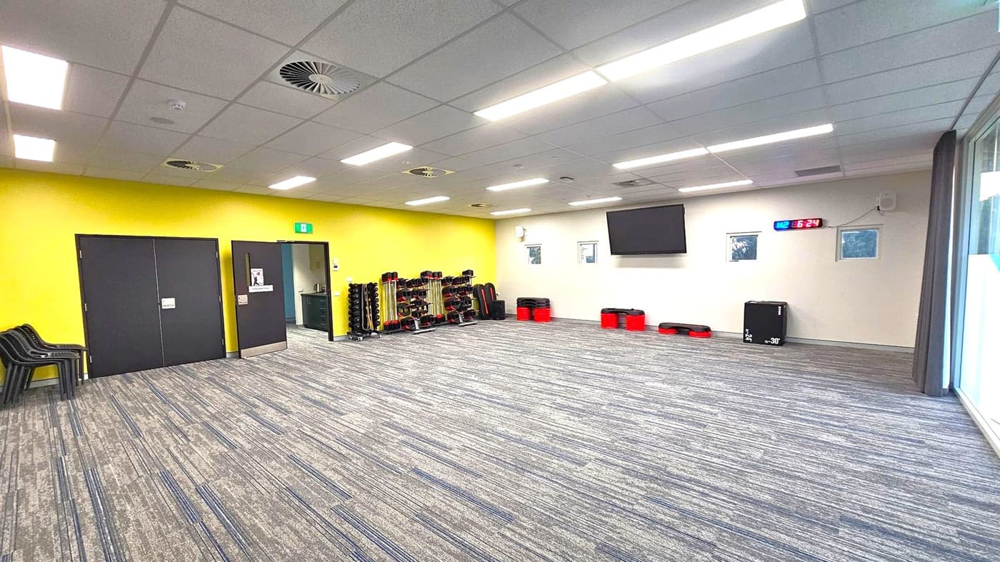 A spacious room with a grey carpeted floor and a ceiling with fluorescent lights and air vents. On the left, there are black double doors and a stack of black chairs. In the middle, a yellow wall features racks of weights and exercise equipment. On the right, a white wall has a large TV screen, a digital clock displaying "16:24", and small windows. Red and black exercise steps and a black box are placed against the wall. Large windows with curtains are on the far right.