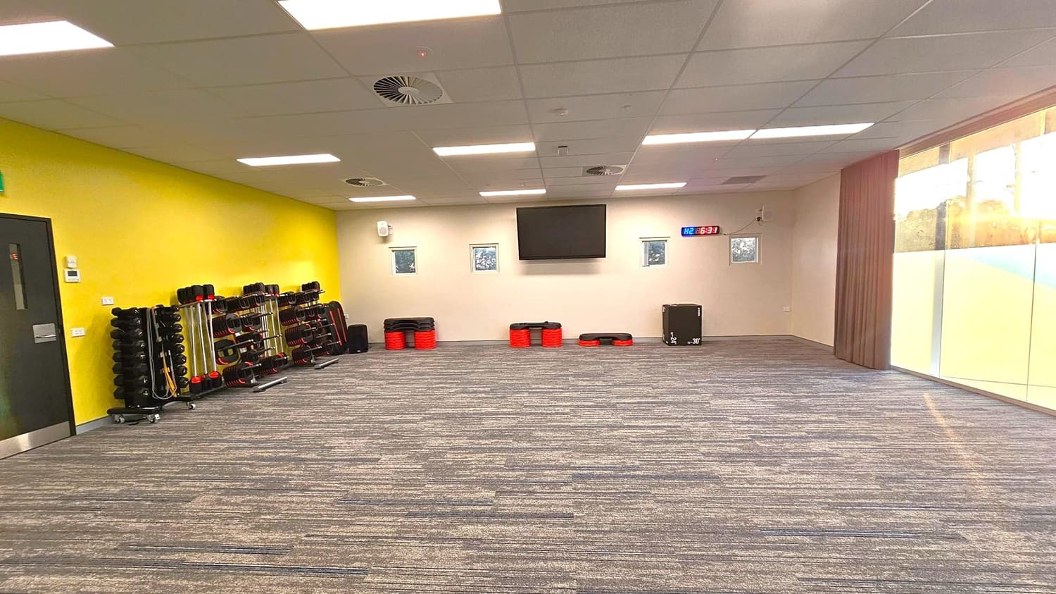 A spacious room with a grey and purple carpeted floor. On the left, a yellow wall features a black door and racks of fitness equipment, including weights and steps. The back wall is white with a large TV screen and a digital clock displaying "6:31". Small windows are positioned on either side of the TV. On the right, large glass windows allow natural light to enter, with a view of trees outside. The ceiling is fitted with white panels and fluorescent lights.