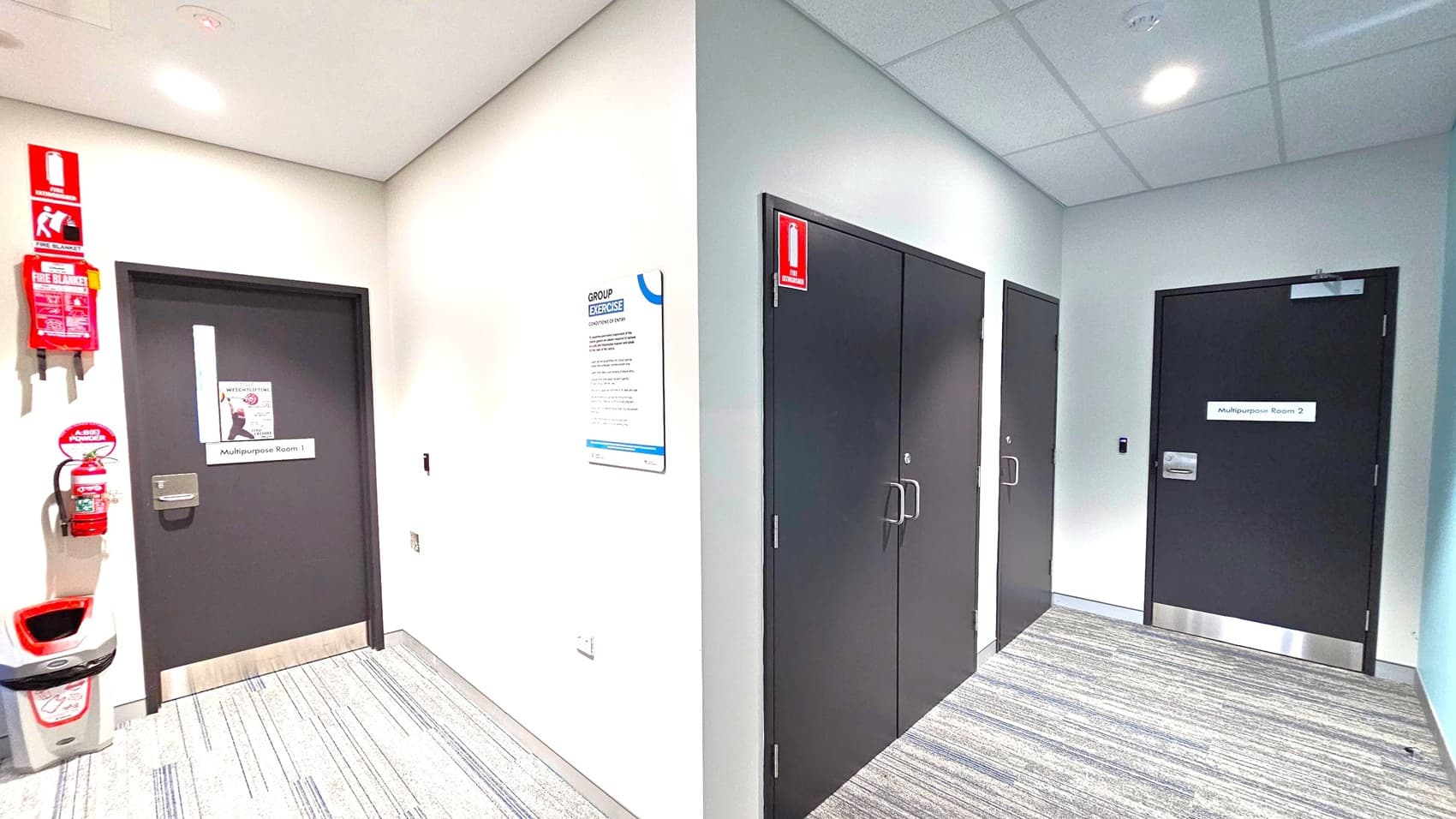 A hallway with grey carpet and white walls. On the left, a door labelled "Multipurpose Room 1" is next to a fire extinguisher and a fire blanket mounted on the wall. A bin is positioned below them. In the centre, a sign titled "Group Exercise" is affixed to the wall. On the right, there are two black doors, one labelled "Multipurpose Room 2". The ceiling is fitted with recessed lighting and a smoke detector.