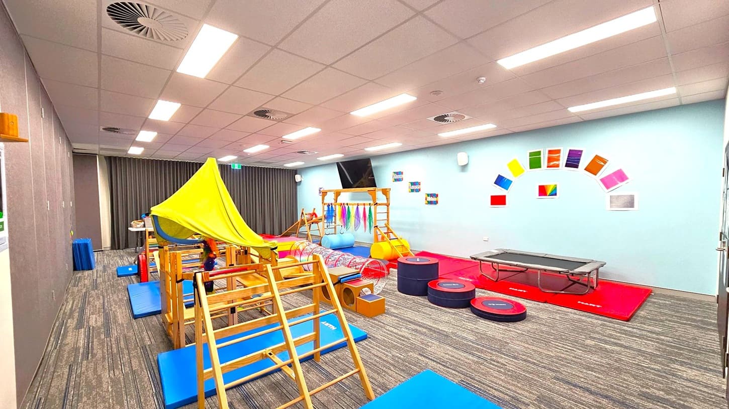 A children's playroom with a variety of equipment. On the left, there's a wooden climbing frame with a green fabric canopy. In the centre, colourful mats and a tunnel are visible. On the right, a trampoline is placed on red mats. The walls are light blue with colourful paper squares arranged in an arc. The floor is carpeted with a striped pattern, and the ceiling has recessed lighting and air vents.