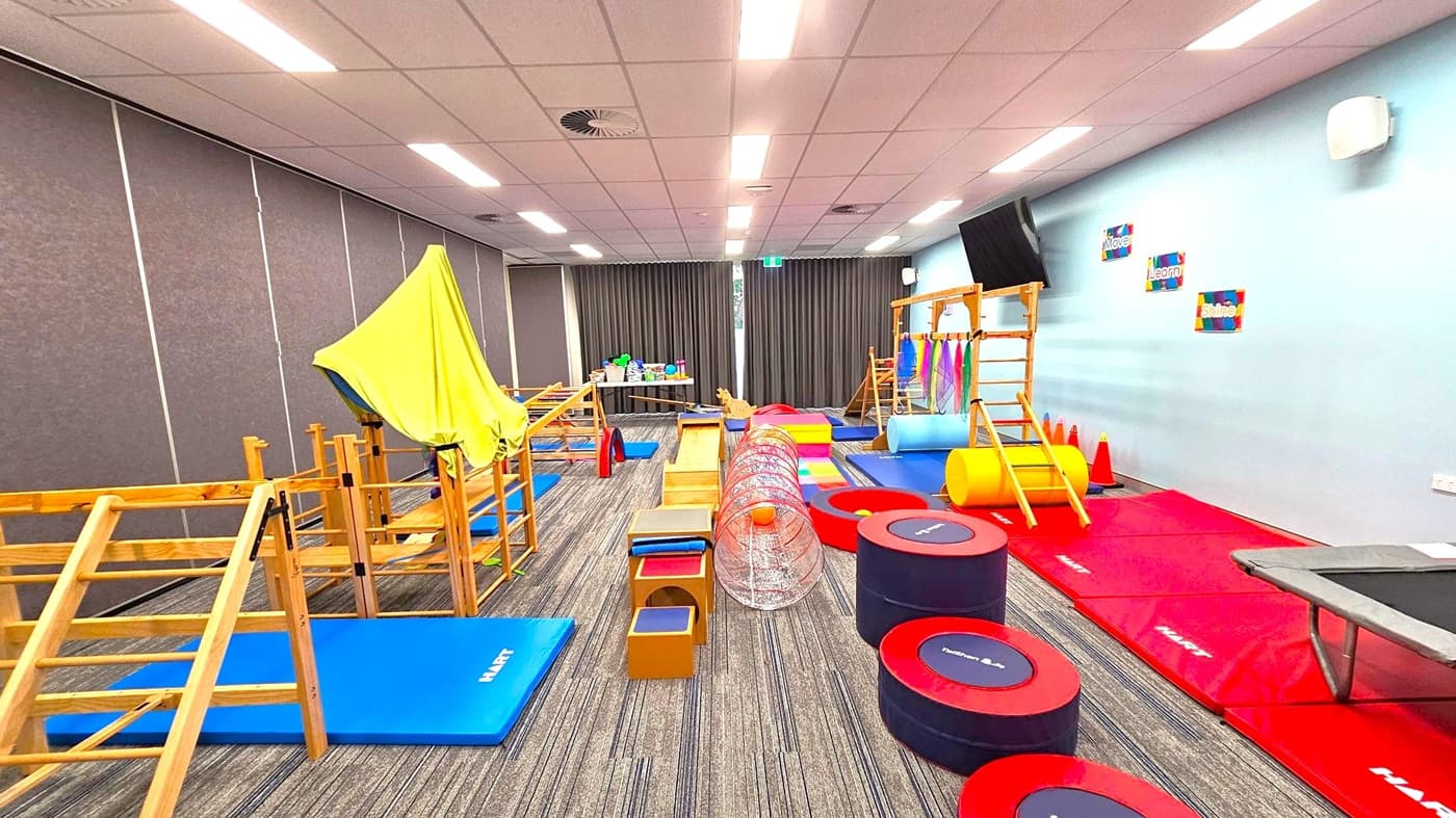 A room with a variety of gym equipment for children. On the left, there are wooden climbing structures and blue mats. In the centre, a tunnel and various soft play items are arranged on a carpeted floor. On the right, red mats, a trampoline, and a wooden ladder are visible. The walls are light blue with motivational posters, and the ceiling has fluorescent lights. A table with toys is at the back near dark curtains.