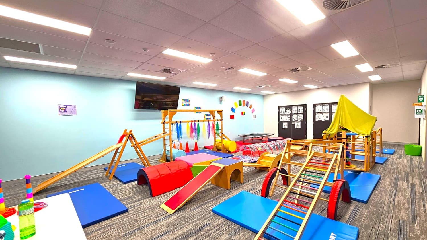 A children's playroom with various wooden climbing structures and colourful mats on the floor. On the left, a table holds stacked toys and bottles. In the centre, a wooden frame with hanging scarves and a slide is surrounded by red and blue mats. On the right, a structure is covered with a green cloth. The walls are light blue with colourful decorations, and a large TV is mounted on the left wall. The ceiling has recessed lighting, and the floor is carpeted with a striped pattern.