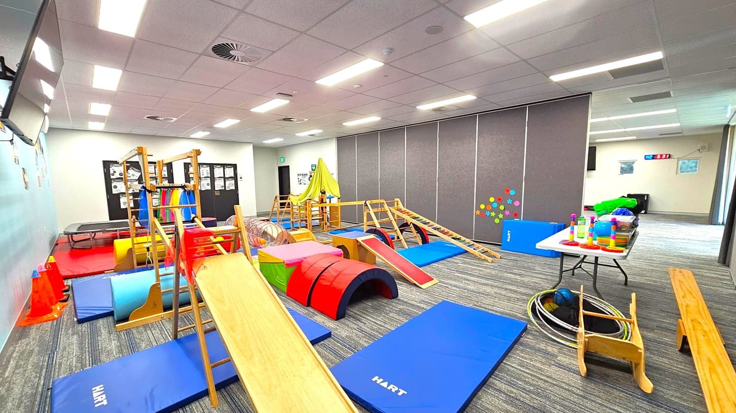 A children's playroom with various gym equipment and toys. On the left, there are wooden climbing structures, colourful mats, and cones. In the centre, a slide and a tunnel are surrounded by soft play shapes. On the right, a table holds stacking toys and other items, with hula hoops and a small wooden chair nearby. The floor is carpeted with a striped pattern, and the ceiling has recessed lighting and ventilation. Walls are decorated with colourful dots and posters.