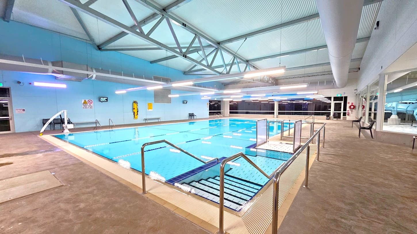 An indoor swimming pool with clear blue water, surrounded by a beige tiled deck. Stainless steel handrails lead into the pool. On the left, a wall with safety signs and a digital clock. The ceiling features exposed metal beams and fluorescent lights. On the right, large windows and glass doors, with a few plastic chairs along the wall. The floor is a textured surface, and the ceiling is lined with ductwork.