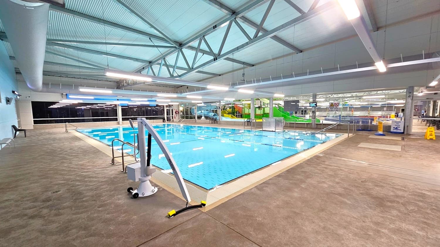 Indoor swimming pool area with a clear blue pool in the centre. The pool is surrounded by a beige tiled floor. On the left, there is a chair against the wall. In the background, large windows and a green water slide are visible. The ceiling is high with exposed beams and fluorescent lights. On the right, there are caution signs and a seating area with tables and chairs.