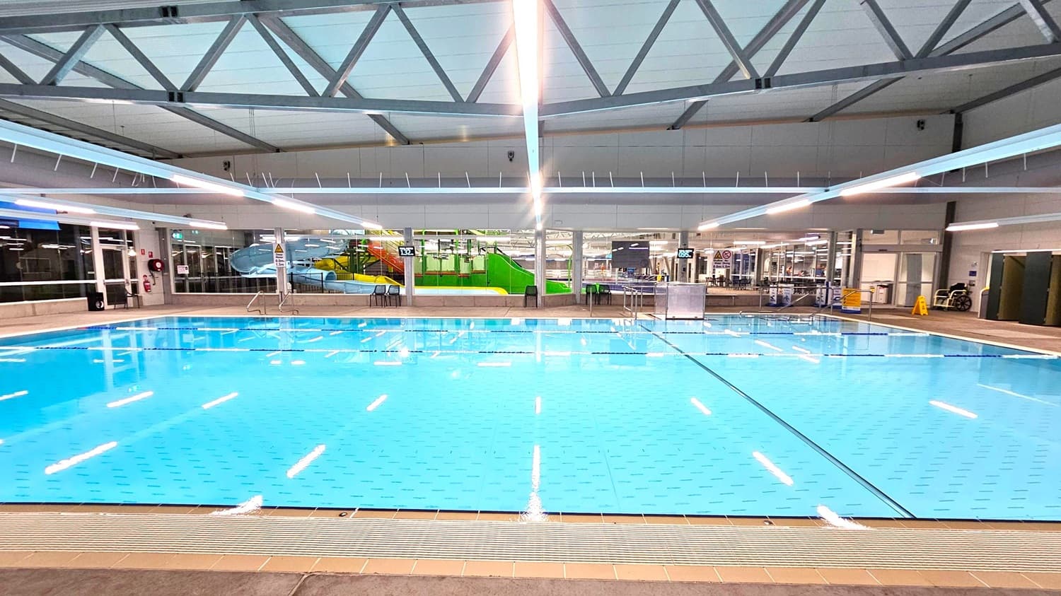 An indoor swimming pool with clear blue water, surrounded by a tiled deck. In the background, there are large windows and a colourful water slide structure with green and yellow elements. The ceiling features exposed metal beams and bright overhead lights. On the right, there are some chairs and a caution sign. The pool area is spacious and well-lit.