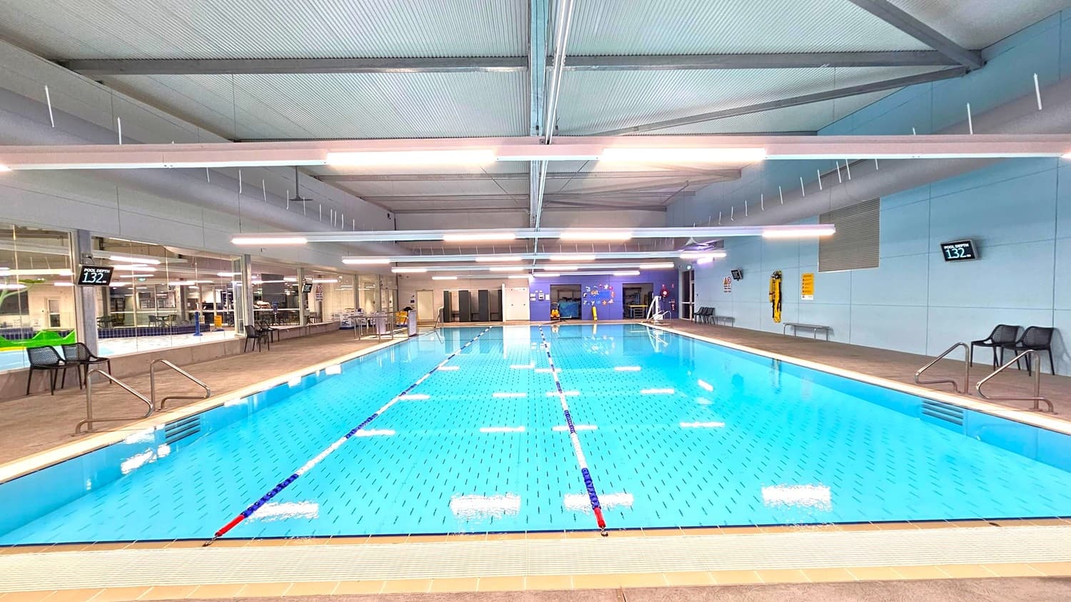 An indoor swimming pool with clear blue water, marked with lane dividers. On the left, there are chairs and a digital display showing the pool depth as 1.32 metres. The right side also has chairs and a similar depth display. The background features large windows, tables, and a wall with safety equipment. The ceiling is high with exposed beams and fluorescent lighting. The floor around the pool is tiled.