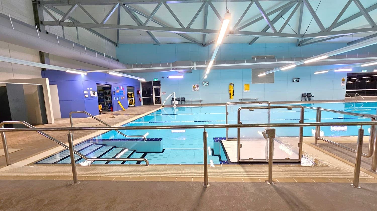 Indoor swimming pool with metal railings in the foreground. Steps lead into the pool on the left, and a hoist is positioned on the right. The pool area is surrounded by a tiled floor. The back wall is blue with various signs and a digital clock. Overhead, there are exposed metal beams and fluorescent lights. A door and some equipment are visible on the left wall.