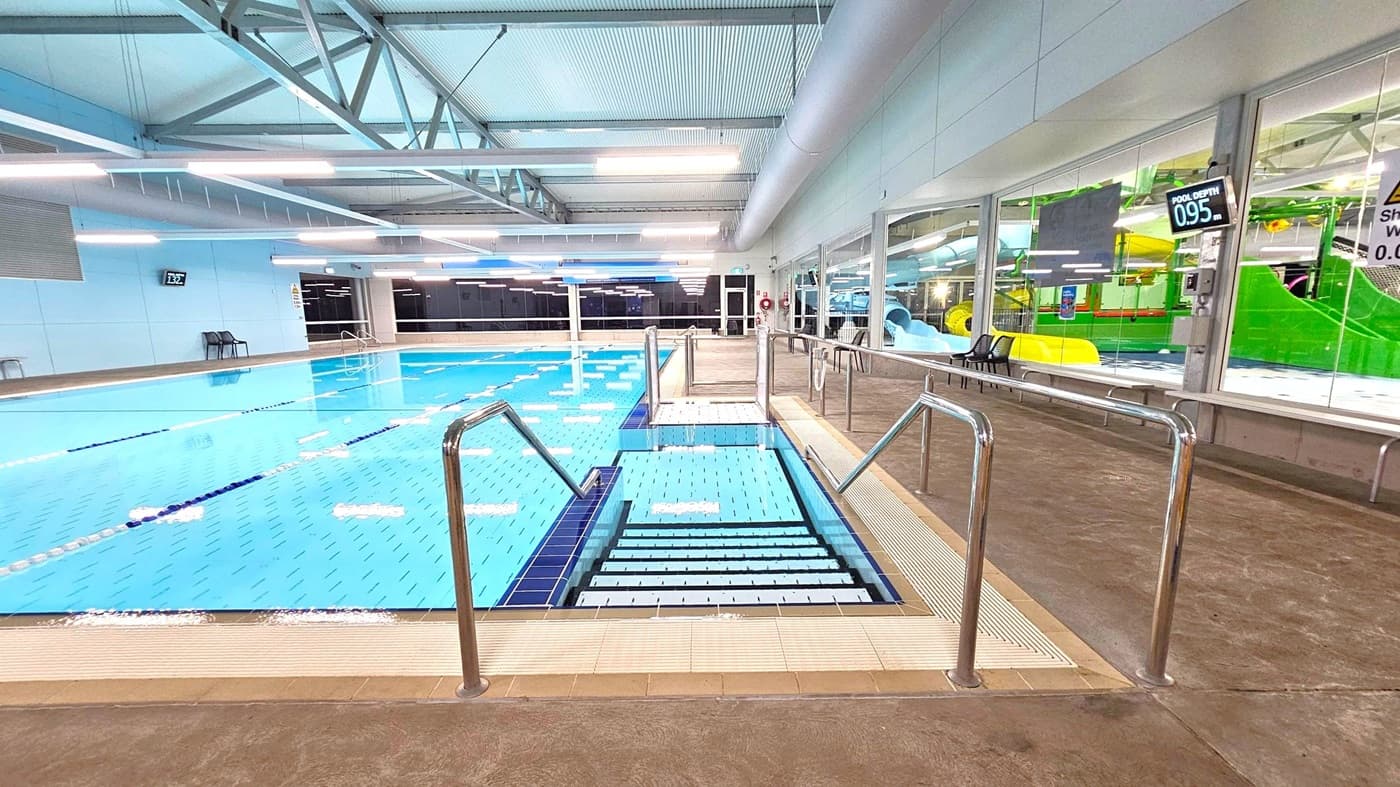 Indoor swimming pool with clear blue water and stainless steel handrails leading into the pool. On the left, the pool extends with lane dividers and a depth sign of 1.32 metres. On the right, a glass wall reveals a play area with colourful slides and structures. The floor is tiled, and the ceiling features exposed beams and bright lighting. A depth sign of 0.95 metres is visible on the right wall.