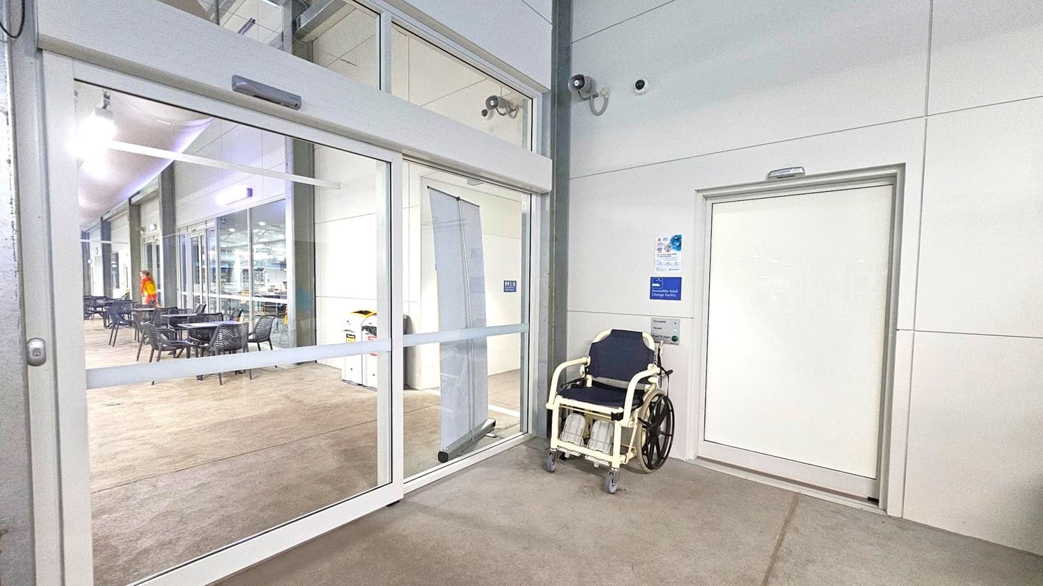On the left, there is a glass sliding door leading to an indoor area with tables and chairs. A person in a red and yellow outfit is visible in the background. On the right, a white door with a sign indicating an accessible adult changing facility is present. A wheelchair is positioned next to the door. The floor is a mix of beige and green, and the ceiling is white with fluorescent lighting. Security cameras are mounted on the wall above the door.