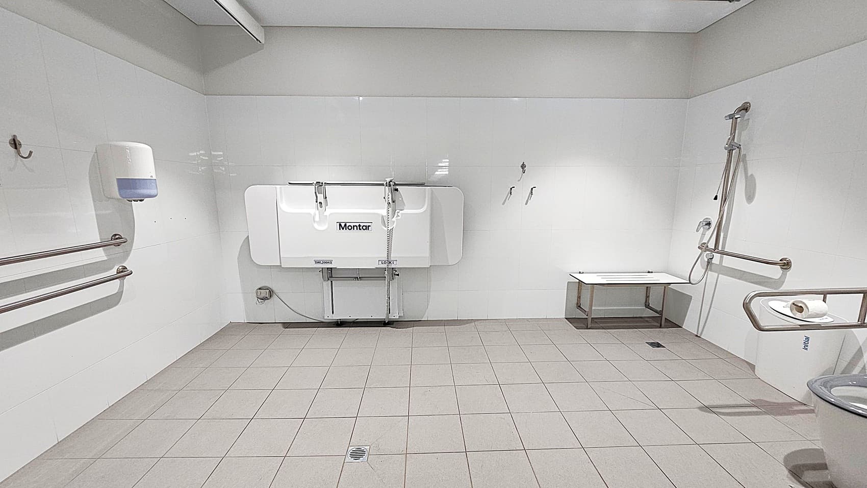 A spacious, accessible bathroom with grey tiled flooring and white walls. On the left, there are horizontal metal grab bars and a wall-mounted soap dispenser. In the centre, a foldable changing table is attached to the wall. On the right, a shower area with a handheld showerhead and a foldable seat is visible. A toilet with a grab bar and a toilet roll holder is in the foreground. The ceiling is plain with a light fixture.