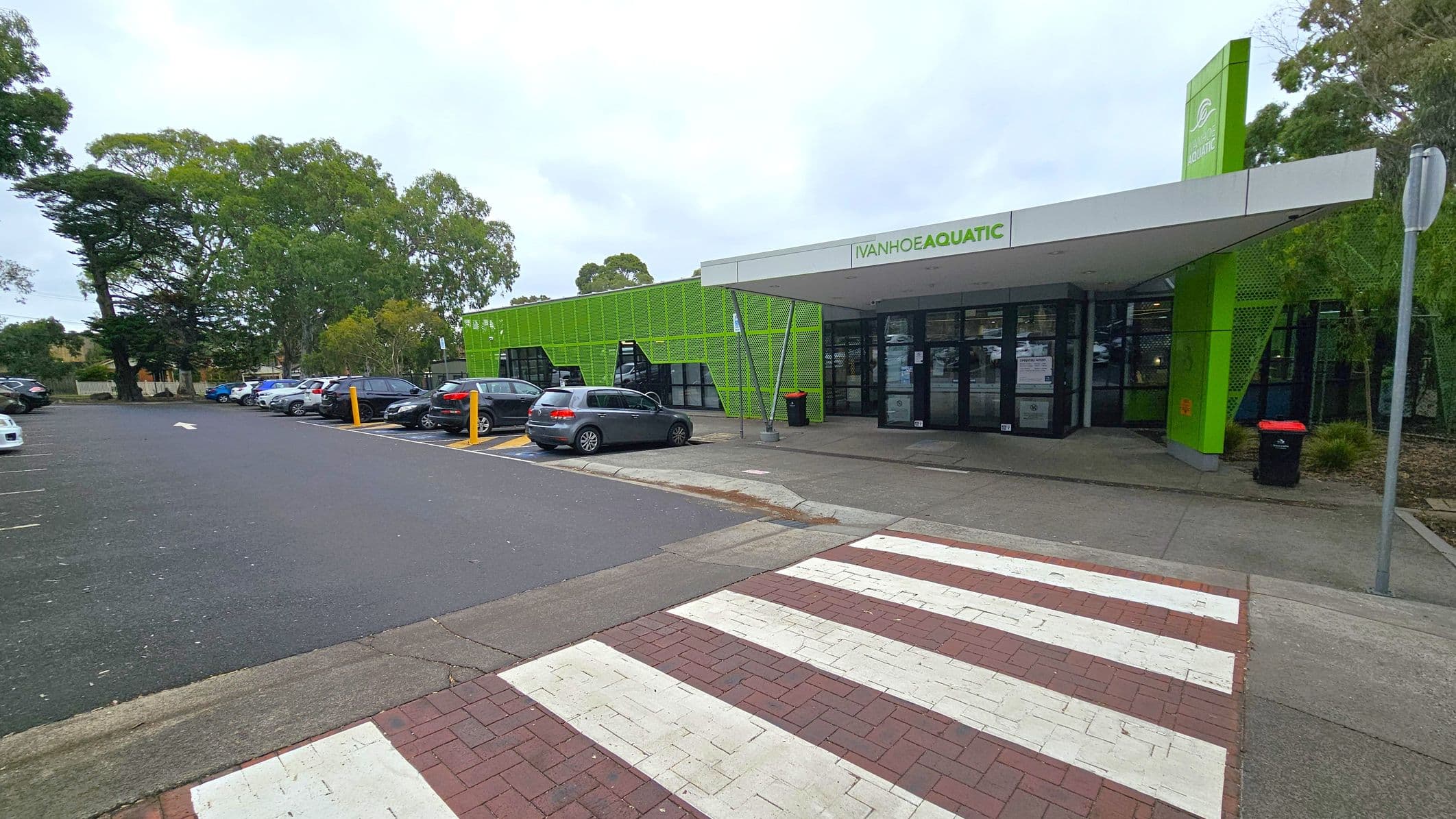 A modern building with a green facade and a sign reading "Ivanhoe Aquatic" above the entrance. The entrance has glass doors and a small awning. To the left, there is a car park with several parked cars and tall trees in the background. A zebra crossing leads to the entrance from the foreground, with a mix of red and white paving. A black bin is near the entrance, and a metal pole stands to the right.