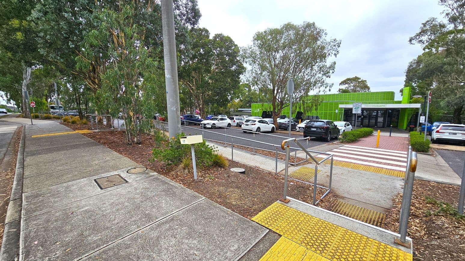A footpath on the left with a concrete surface and a yellow tactile paving section leads to a road. On the right, a car park with several vehicles is visible, adjacent to a green building with a sign that reads "AQUATIC." Trees are scattered throughout the area, providing shade. A pedestrian crossing with red and white stripes connects the footpath to the building entrance. A metal railing and a utility pole are also present near the crossing.