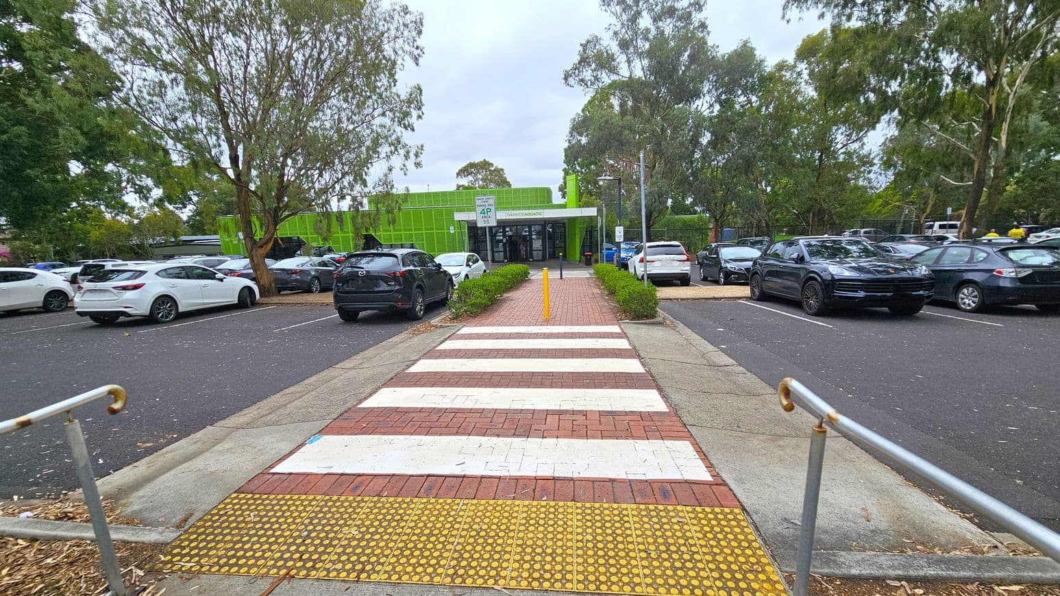 A pathway with a zebra crossing leads towards a building with a bright green facade in the centre. On either side of the path, there are parked cars, including white and dark-coloured vehicles. Trees with dense foliage are visible on both sides, providing shade. The ground is paved with bricks and asphalt, and there are metal railings on the left. The building has a sign above the entrance, and the area appears to be a car park.