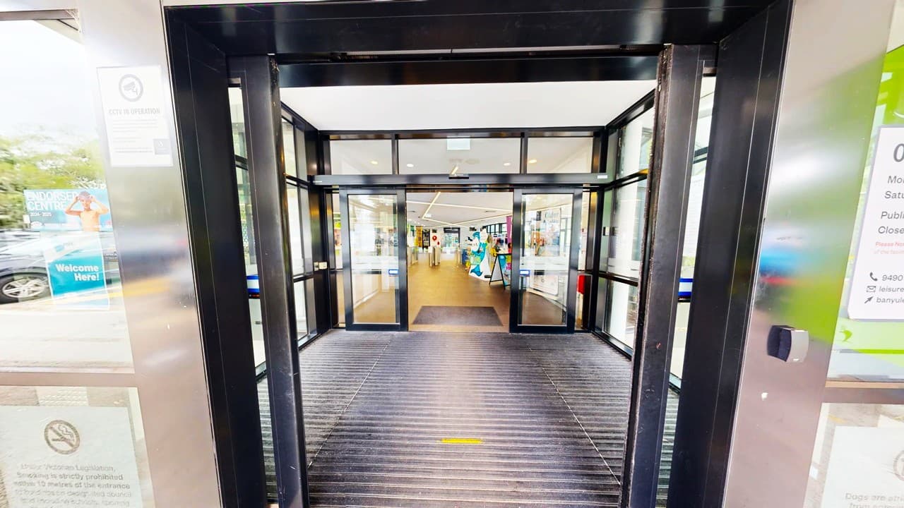 Automatic sliding glass doors lead into a building with a bright interior. The entrance has a black frame and a ribbed mat on the floor. Inside, the floor is wooden, and there are displays and signs visible in the distance. On the left, there are posters and signs on the glass, while the right side also features signage. The ceiling inside is white with recessed lighting.