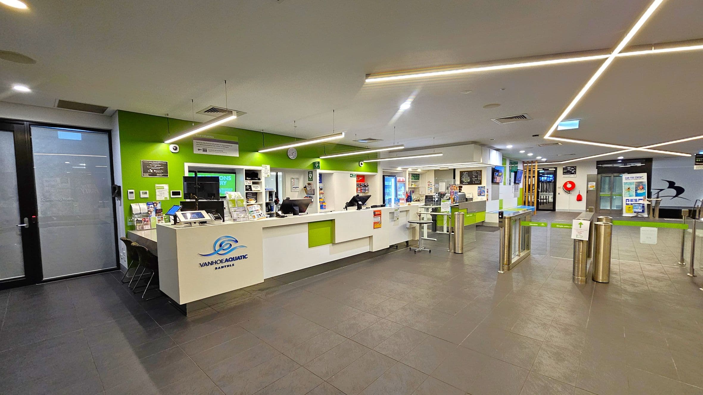 A modern reception area with a green and white colour scheme. On the left, there's a counter with brochures and a computer, and a door with frosted glass. In the centre, the reception desk displays the "Ivanhoe Aquatic" logo, with computers and informational materials. On the right, there are turnstiles leading to another area. The ceiling features linear lights, and the floor is tiled in dark grey. Various signs and posters are visible throughout the space.