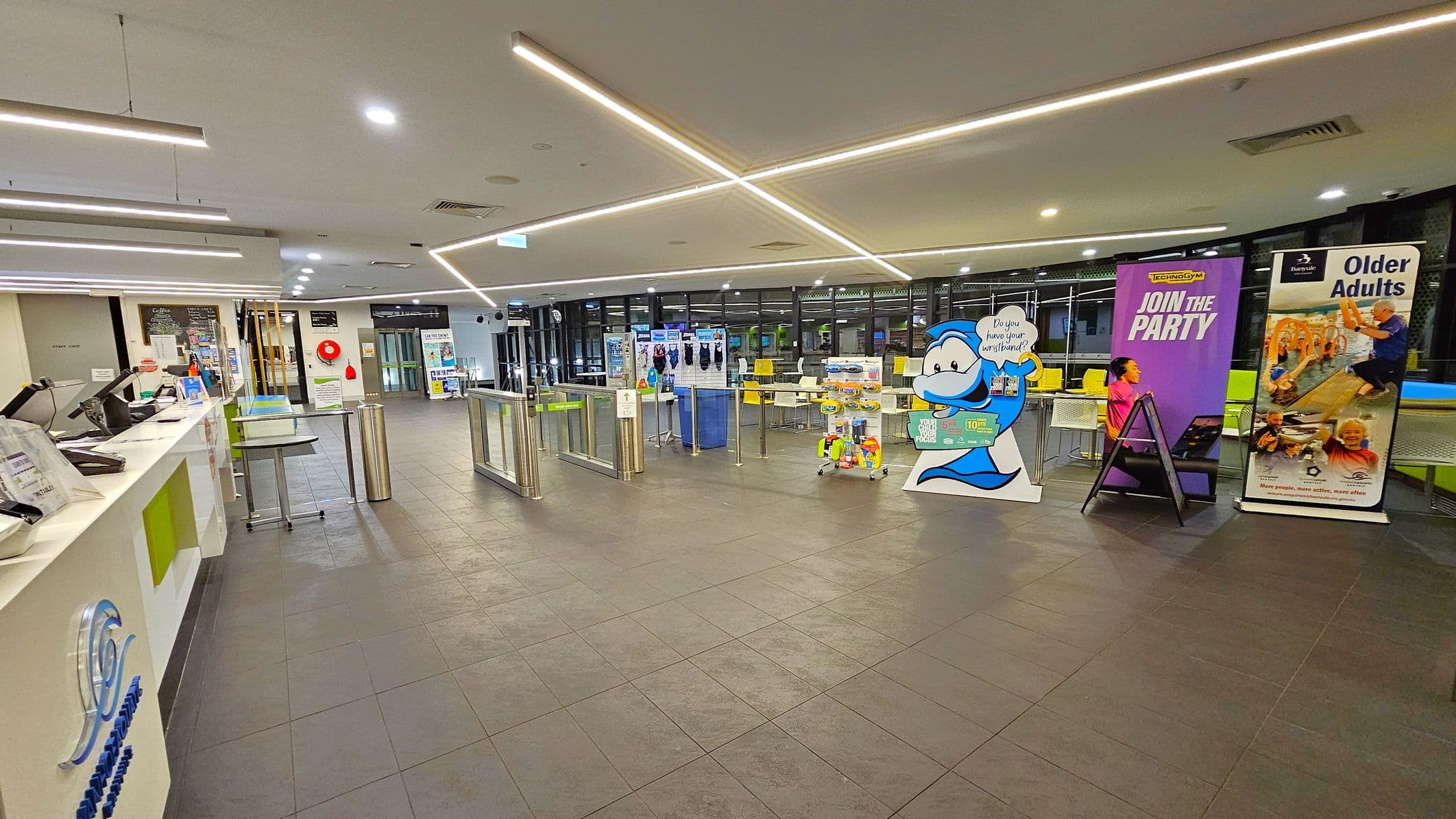 A spacious indoor area with a tiled floor and a ceiling featuring linear lights. On the left, there's a reception counter with computers and informational materials. In the centre, there are turnstiles leading to a seating area with tables and chairs. On the right, several promotional banners are displayed, including one with a cartoon character and others advertising activities for adults. The background has large windows and additional seating.