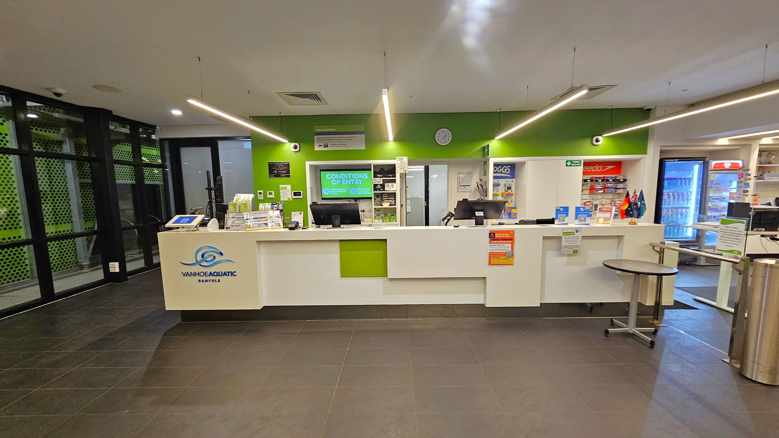 A reception area with a white counter featuring a green accent and a logo. Behind the counter, a green wall displays a clock and various signs. To the left, there are large windows with a perforated green pattern. On the right, shelves with products and a refrigerator with drinks are visible. The floor is tiled in dark grey, and the ceiling has linear lights. A small round table is in front of the counter.