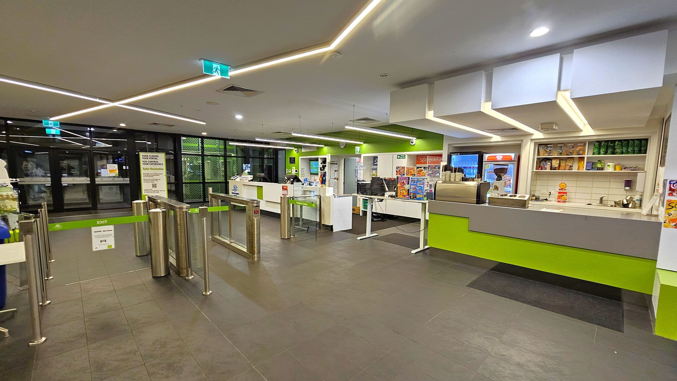 A modern indoor space with a tiled floor and a ceiling featuring linear lights. On the left, there are glass doors with a green exit sign above and a turnstile gate with a green exit barrier. In the centre, a counter with various items and a sign. On the right, a service counter with a coffee machine, shelves stocked with snacks, and a menu board. The walls are painted green and white, and the area is well-lit.