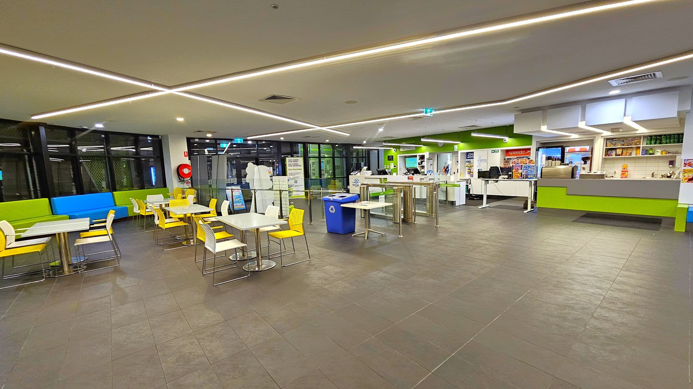 A spacious indoor cafeteria with a tiled floor and a ceiling featuring linear lights. On the left, there are tables with white tops and yellow chairs, alongside green and blue cushioned bench seating. In the centre, a blue recycling bin is visible. On the right, a counter area with green accents displays various items, with shelves holding products above. The background includes large windows and a fire extinguisher mounted on the wall.