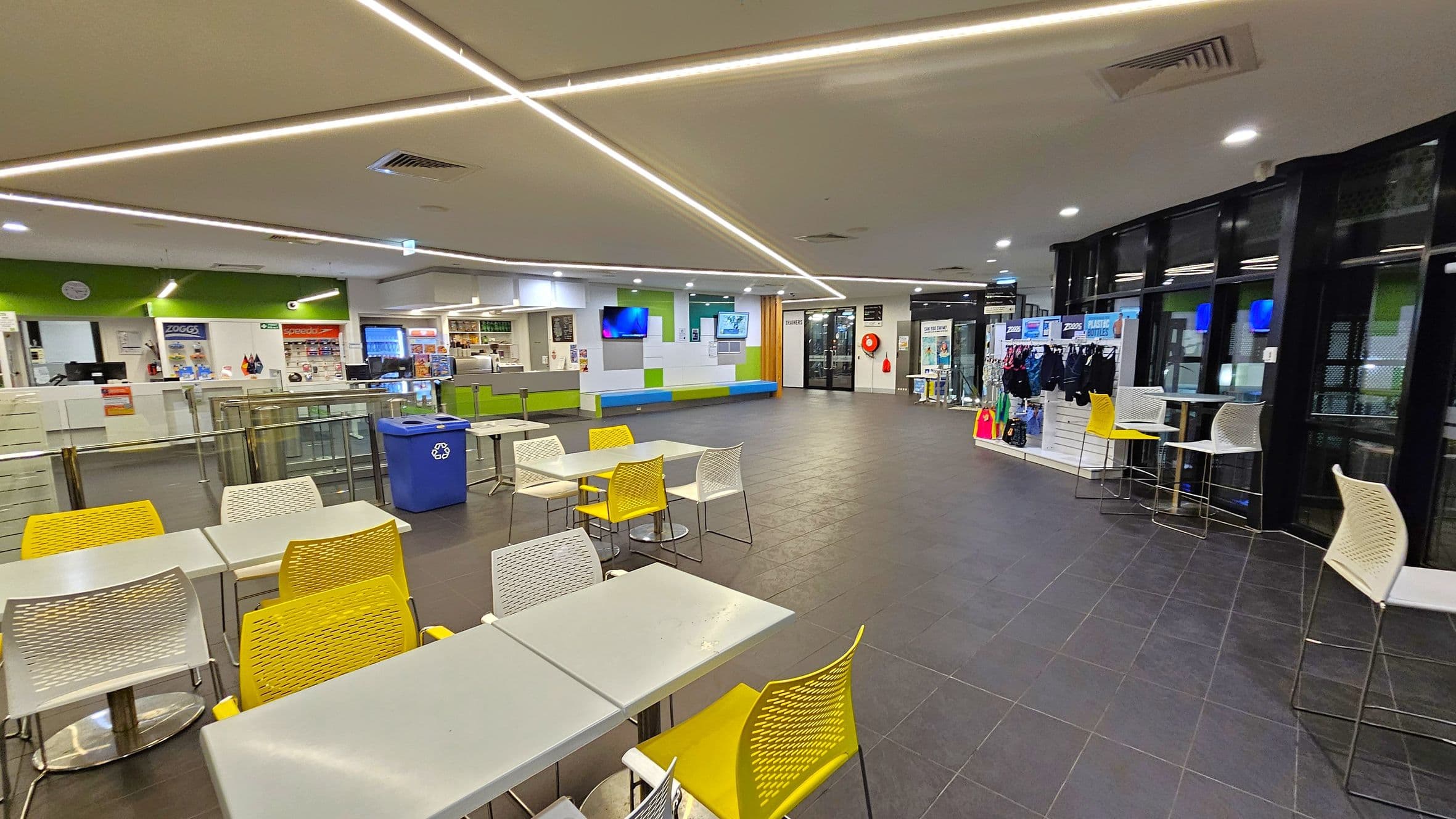 A spacious indoor area with a tiled floor and a ceiling featuring linear lights. On the left, there are tables with yellow and white chairs, and a blue recycling bin. The background has a counter with a green accent wall, a clock, and a display of products. In the centre, a wall-mounted TV and a seating area with green and blue cushions are visible. On the right, there is a display of swimwear and accessories, with high tables and chairs near large windows.