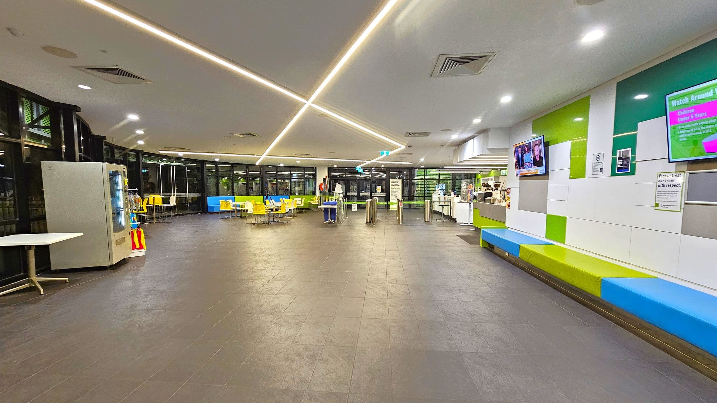 A spacious indoor area with a dark tiled floor and a ceiling featuring recessed lighting and linear light strips. On the left, there's a vending machine and a small table. In the centre, yellow chairs and tables are arranged near large windows. On the right, a wall with colourful panels has two mounted screens and a long bench with blue and green cushions. A counter with various items is visible further back on the right.