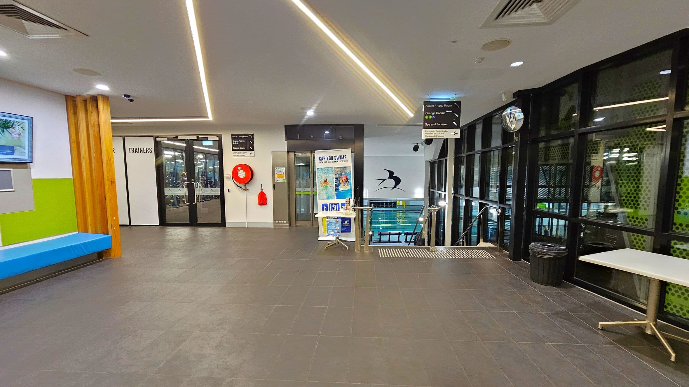 A modern indoor facility with a grey tiled floor and a white ceiling featuring recessed lighting. On the left, there's a blue bench against a wall with green and white sections, and a digital screen above. In the centre, glass doors labelled "TRAINERS" lead to another room. A red fire hose reel is mounted on the wall. To the right, large windows overlook a pool area, with a table and a bin nearby. A sign above directs to various amenities, and a small table with posters stands near the pool entrance.