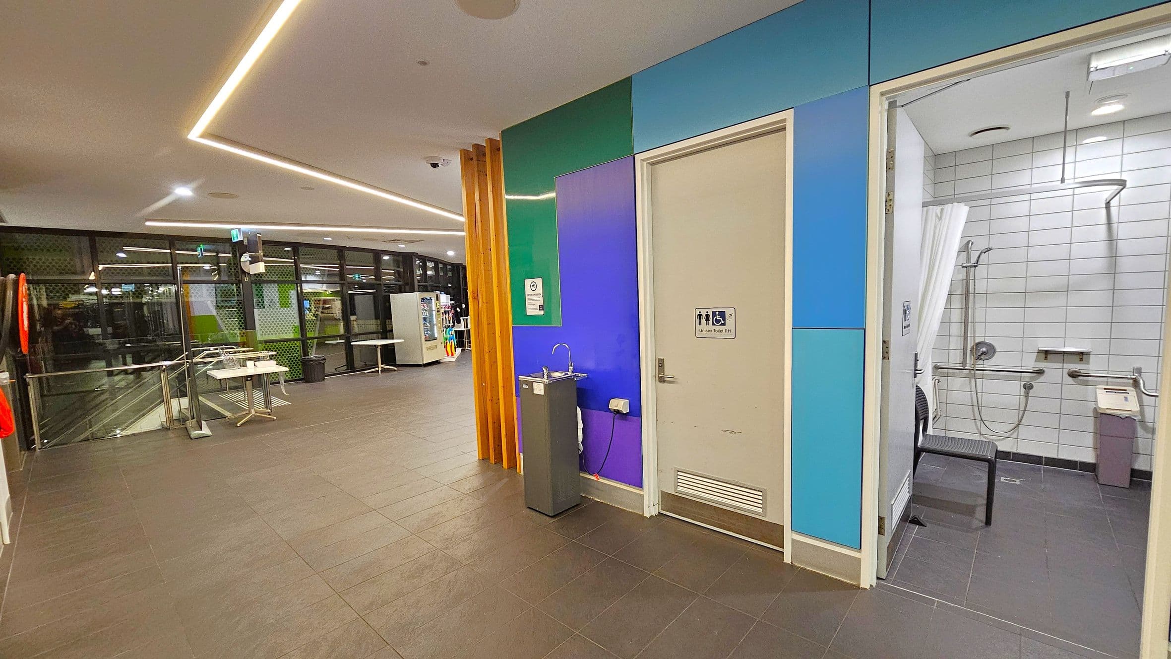 A modern indoor space with a tiled floor and a ceiling featuring recessed lighting. On the right, there is an accessible bathroom with white tiled walls, a shower, and a chair. The door is surrounded by panels in shades of blue and green. A drinking fountain is positioned next to the door. On the left, large windows with a view of the outside are visible, along with a vending machine and tables. A life preserver is mounted on the wall near the windows.