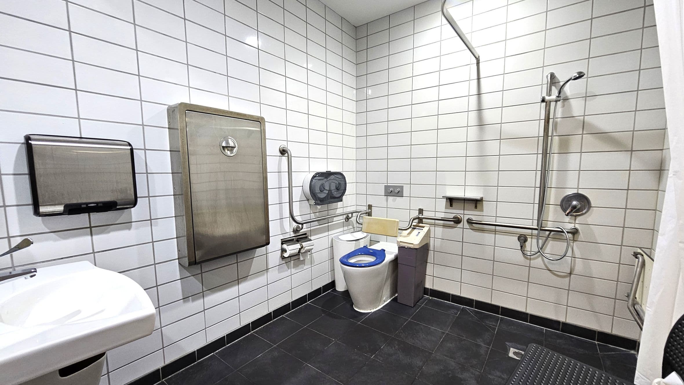 A bathroom with white tiled walls and a dark tiled floor. On the left, there's a white sink with a soap dispenser and a paper towel holder above it. In the centre, a toilet with a blue seat is equipped with support rails and a sanitary bin nearby. On the right, a shower area features a handheld showerhead and additional support rails. A large stainless steel panel is mounted on the wall above the toilet. The ceiling is plain and white.