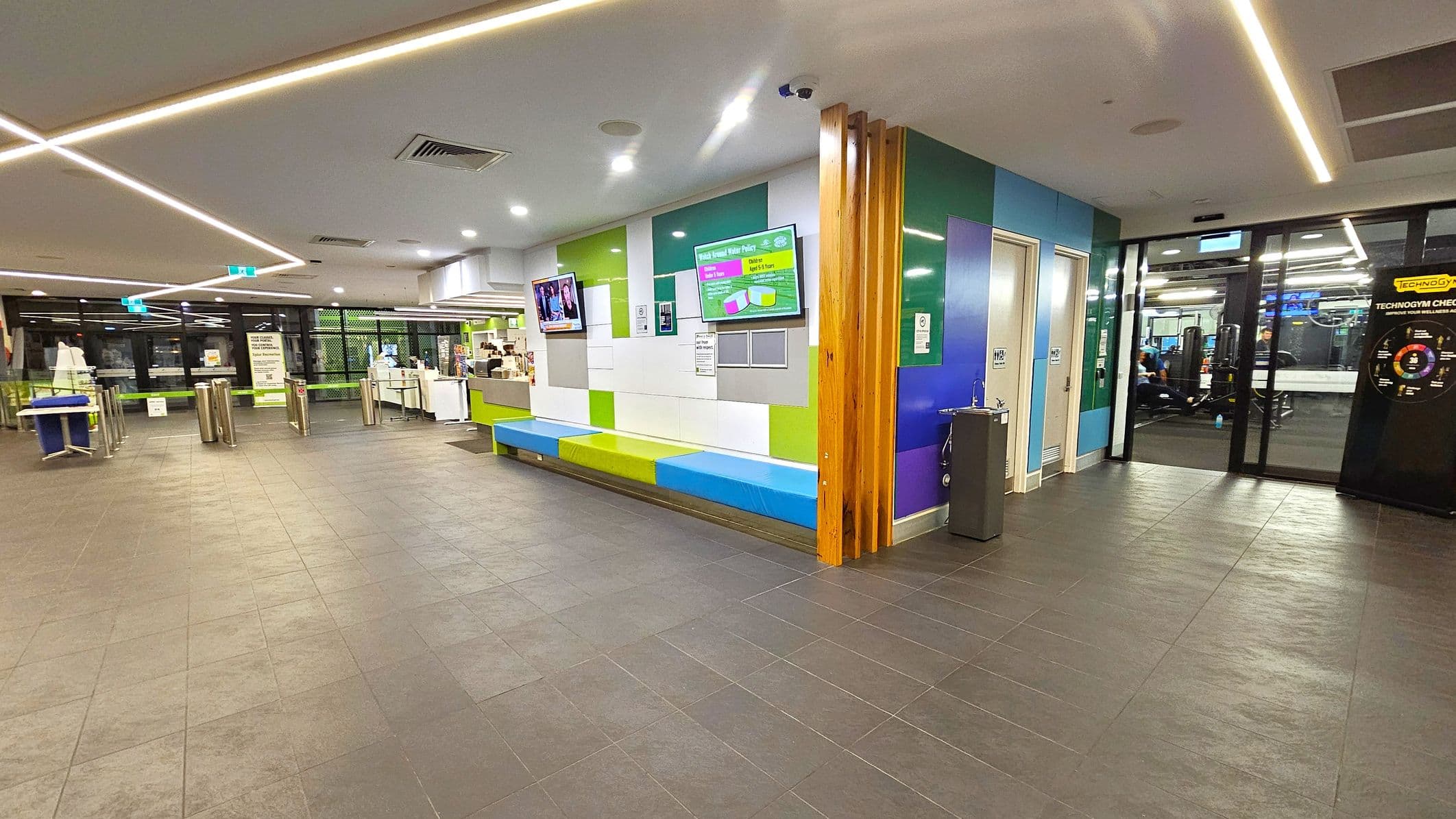 A modern interior with a tiled floor and a ceiling featuring recessed lighting. On the right, there is a colourful wall with a bench and digital screens displaying information. A water fountain is nearby. On the left, there are turnstiles and a reception area with a counter. Glass doors lead to a gym area with exercise equipment visible. The space is well-lit and has a contemporary design.